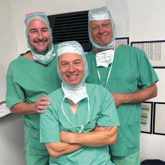 Drei Ärzte in grüner OP-Kleidung stehen gemeinsam im Klinikraum und lächeln in die Kamera.