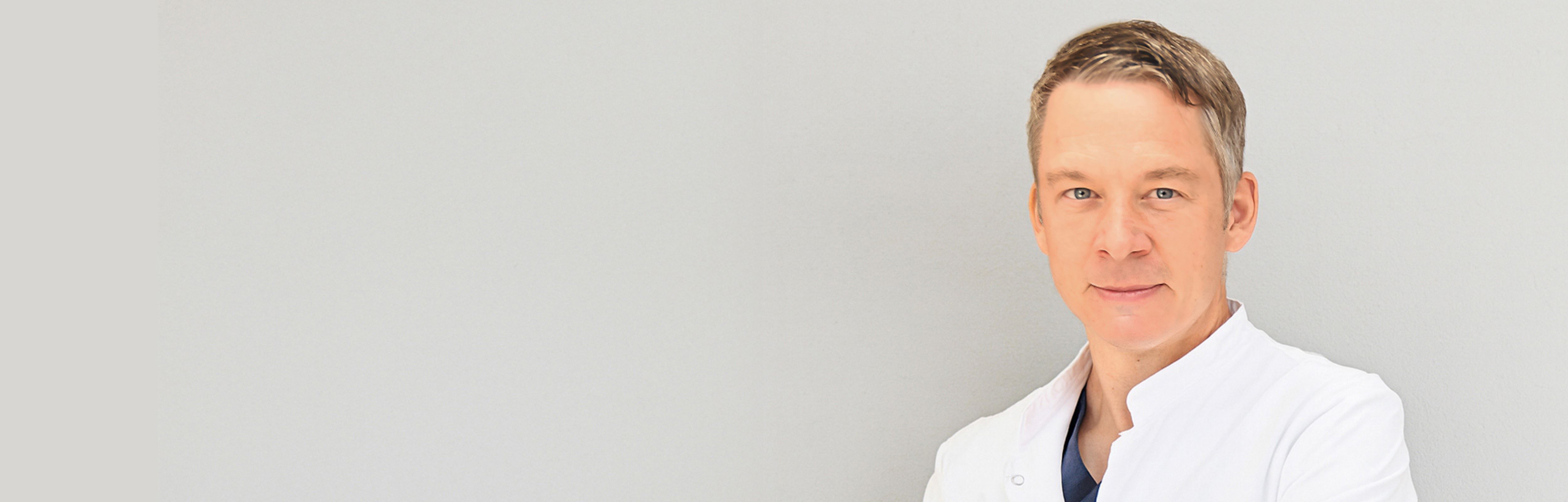 Dr. med. Andreas Bell im weißen Arztkittel, Arme verschränkt.