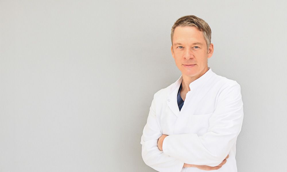 Dr. med. Andreas Bell im weißen Arztkittel, Arme verschränkt.