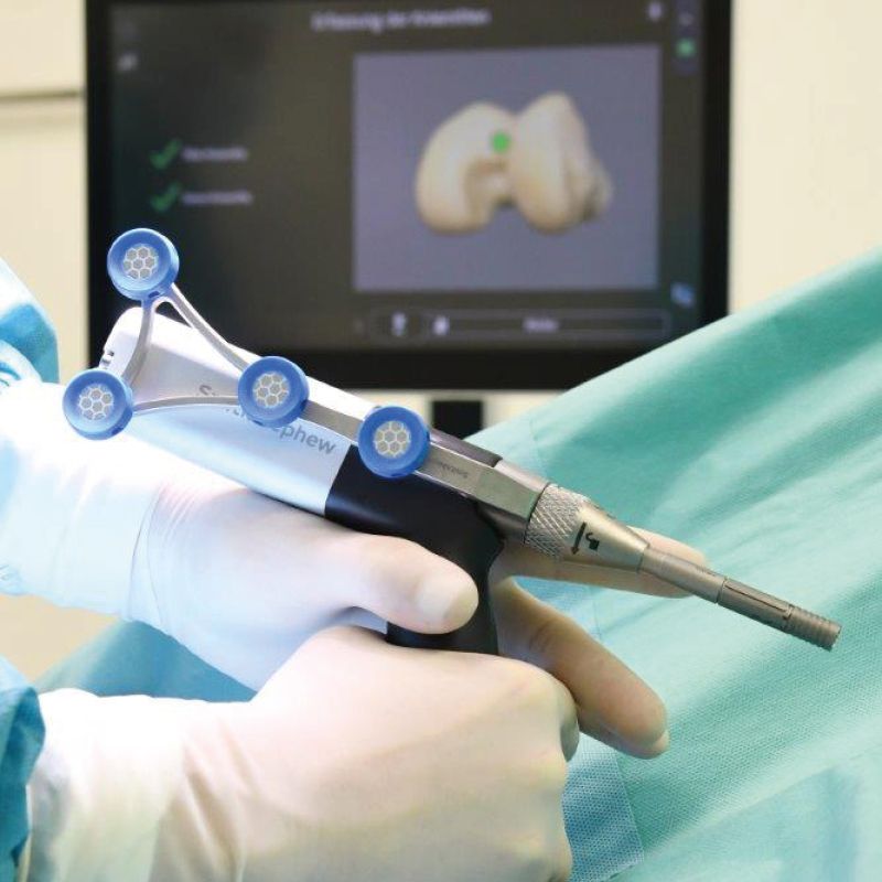 Chirurg hält ein robotergestütztes Instrument zur Vorbereitung einer Knieendoprothese, im Hintergrund ein Monitor mit 3D-Kniemodell.