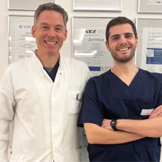 Zwei Ärzte stehen nebeneinander vor einer Wand mit gerahmten Zertifikaten.