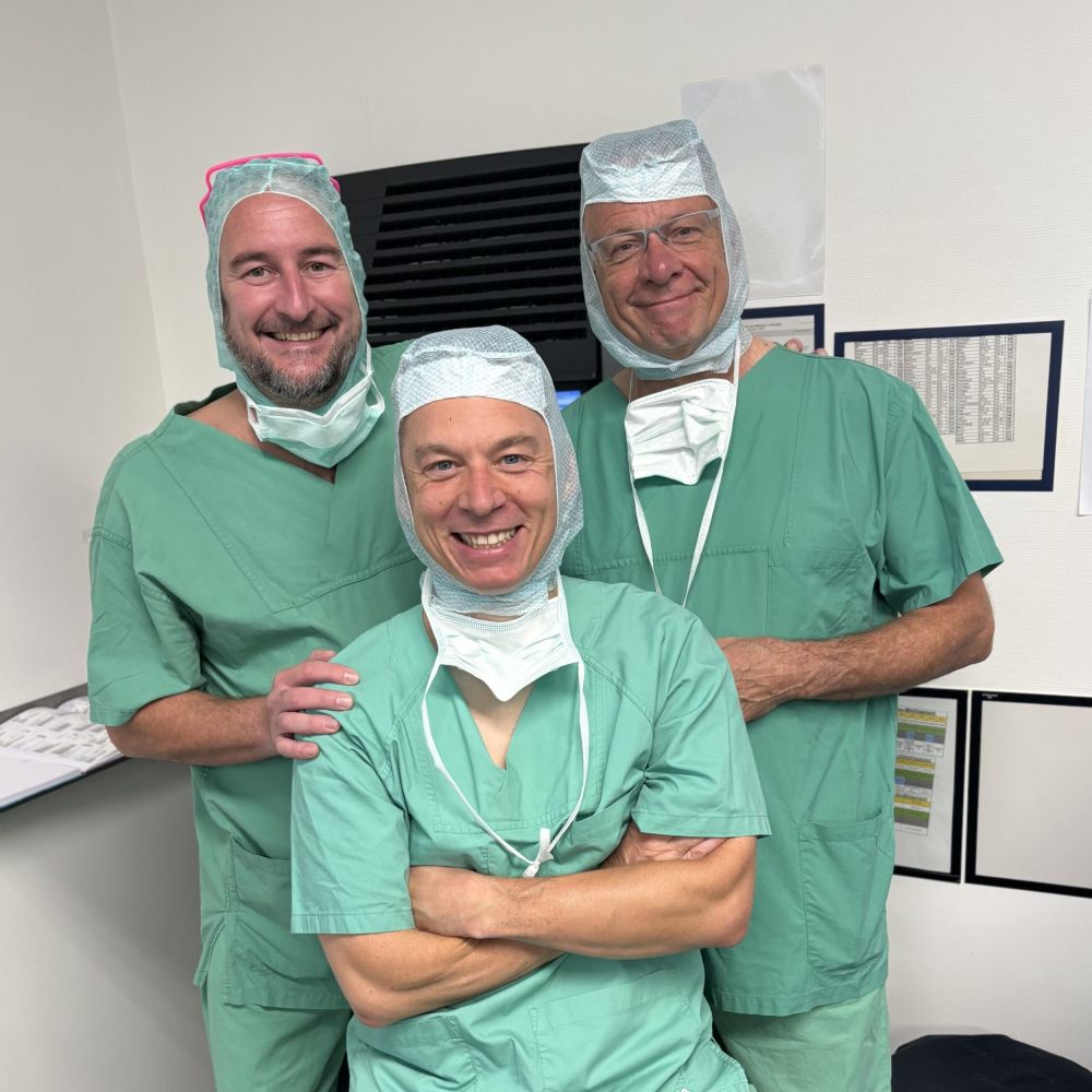 Drei Ärzte in grüner OP-Kleidung stehen gemeinsam im Klinikraum und lächeln in die Kamera.