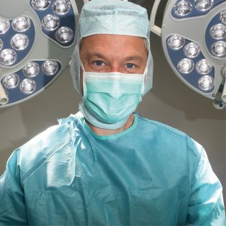 Dr. med. Andreas Bell bei einer OP in grünem OP Kittel mit OP Lampen im Hintergrund.